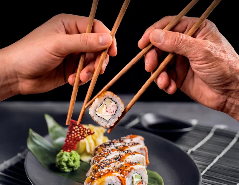 Duas mãos com hashi disputando um uramaki sobre um prato escuro.