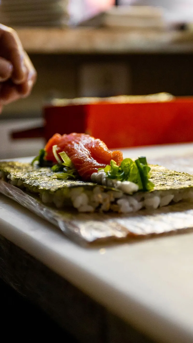 As mãos do sushiman montando um enrolado de salmão sobre a alga, na tábua do balcão.